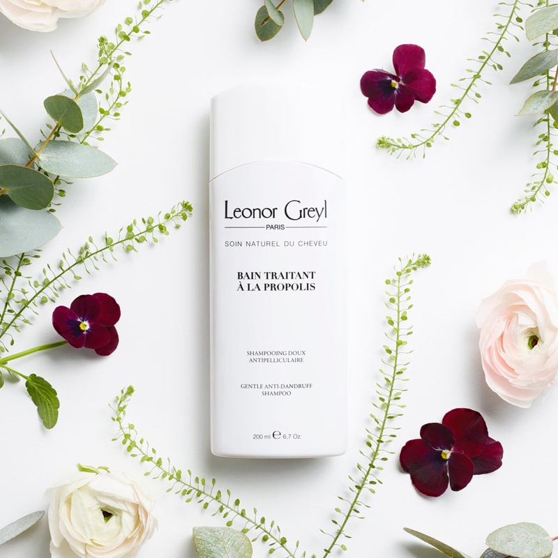 Lifestyle shot top view of Leonor Greyl Bain Traitant A La Propolis – Gentle Dandruff Treatment Shampoo with pink and burgundy flowers and green leaves in the background