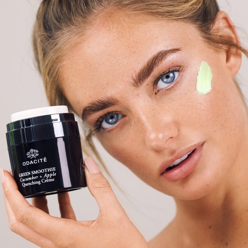 Model holding jar of Odacite Green Smoothie Quenching Creme and showing smear on face