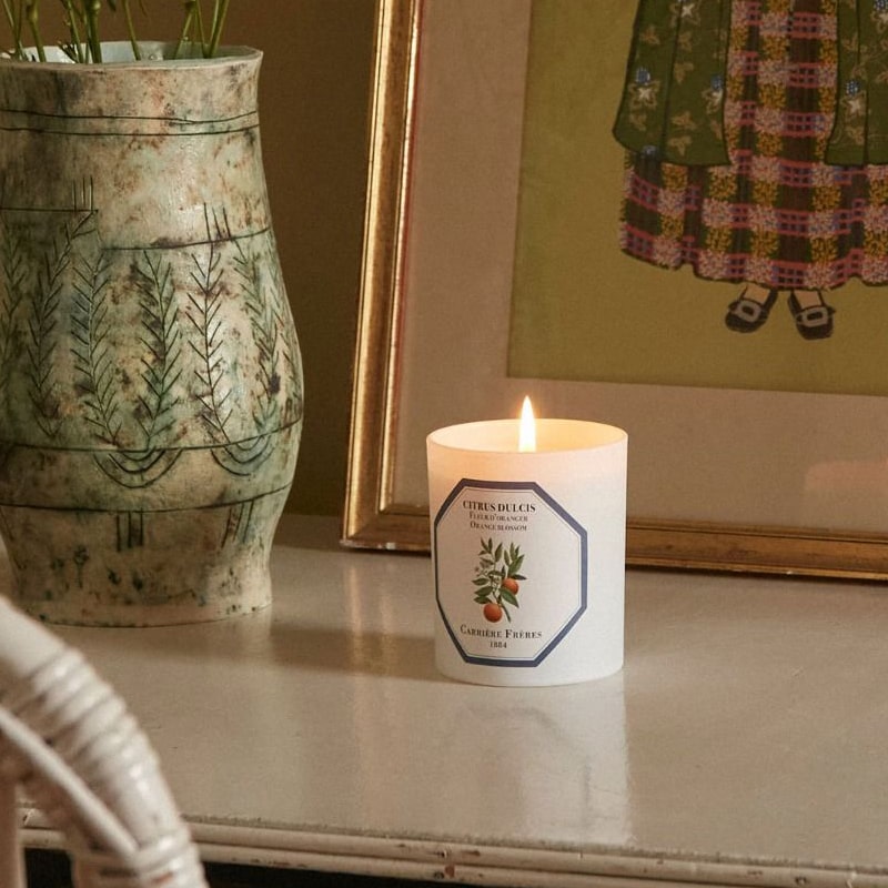 Lifestyle shot of Carriere Freres Orange Blossom Candle showing lit candle next to vase and framed art in the background