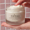 A hand holds a clear jar of Rahua Leave-In Treatment (60 ml) showing the white cream being scooped with a finger, set against a pink tiled background