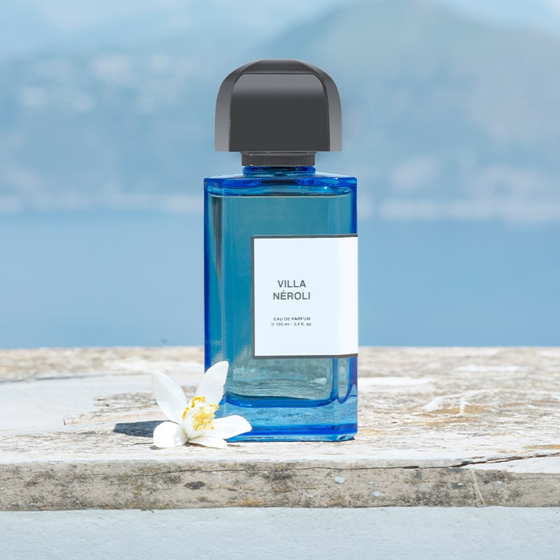 BDK Parfums Villa Neroli Eau de Parfum - bottle sitting on stone slab next to flower in front of mountains and ocean