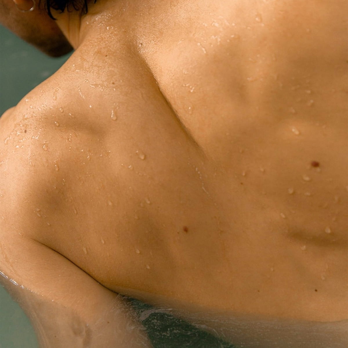 Close-up of a person's bare upper back and shoulder with water droplets on the skin, partially submerged in water using Susanne Kaufmann Bath for the Senses