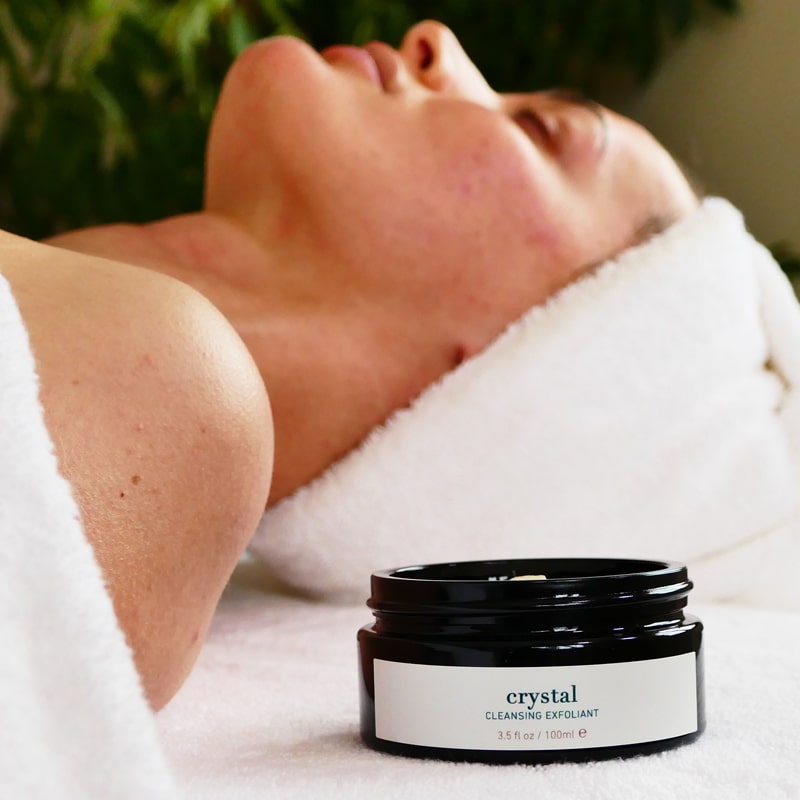 Lifestyle shot of ISUN Crystal Cleansing Exfoliant (100 ml) in the foreground and woman awaiting facial treatment in the background