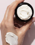 Close up of model holding opened jar of Odacite Jojoba Pearls Daily Hydra-Exfoliant (50 ml) and product smear on hand