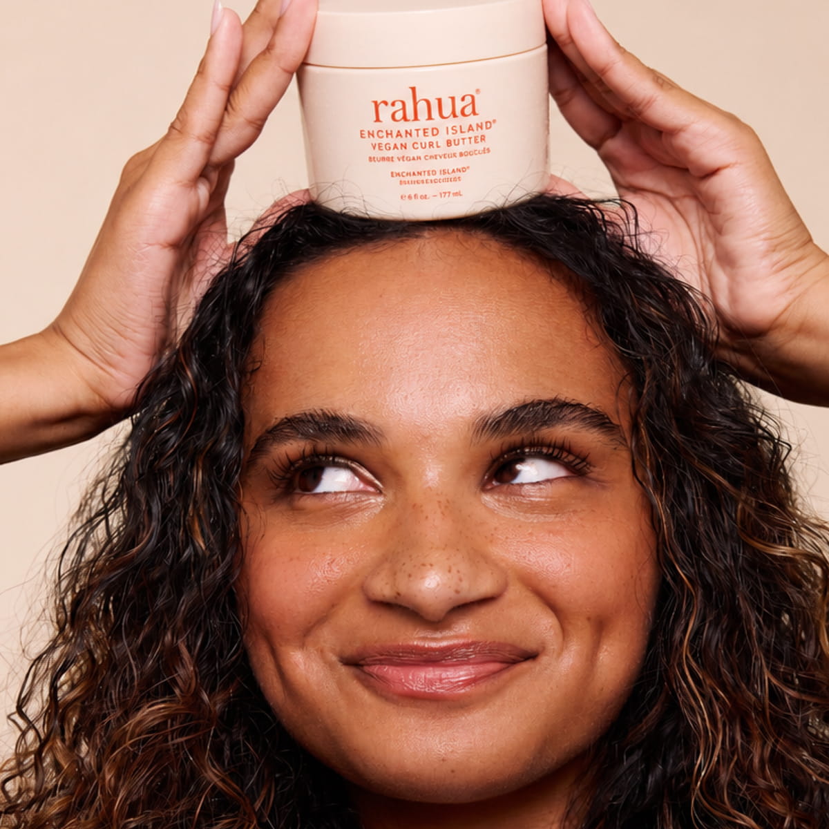 A woman with curly dark hair holds a jar of Rahua Enchanted Island Vegan Curl Butter (177 ml) above her head
