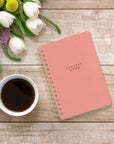 Studio Oh! Agatha Notebooks - Current Mood Coral Pink - lifestyle shot of notebook, pen, coffee cup, and flowers on wood table 