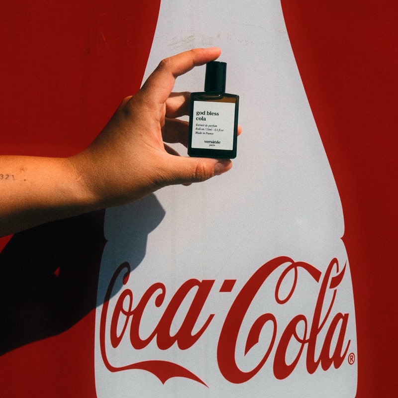 Versatile Paris God Bless Cola Extrait de Parfum in model's hand in front of a Coca-Cola sign.