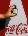 Versatile Paris God Bless Cola Extrait de Parfum in model's hand in front of a Coca-Cola sign.