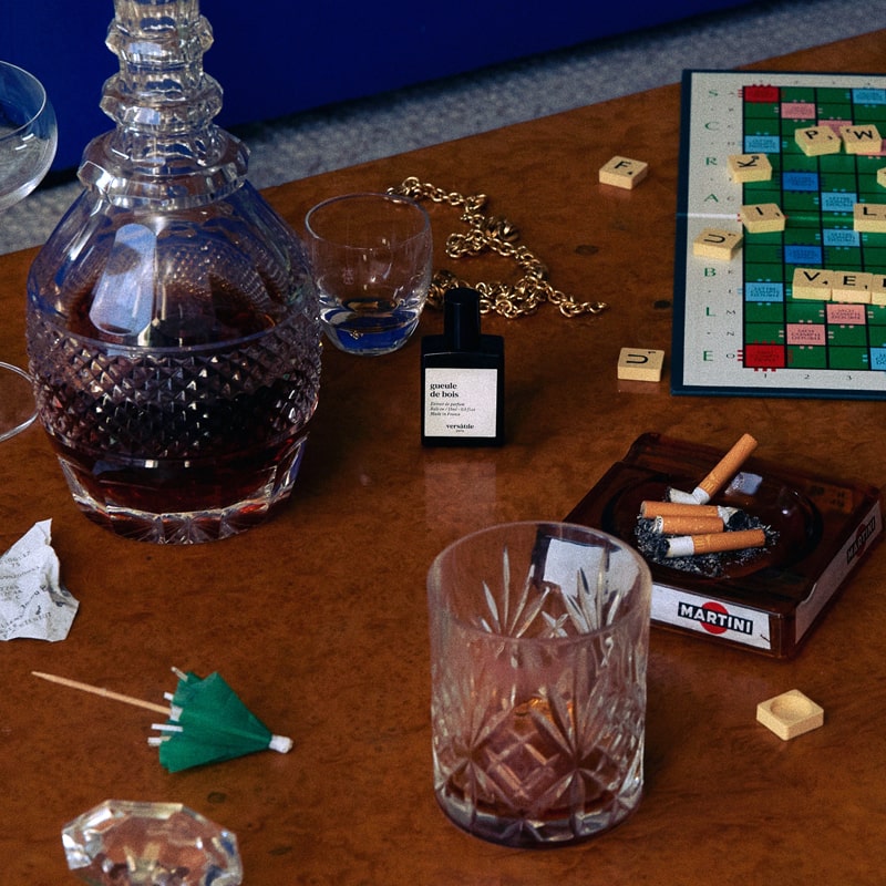 Versatile Paris Gueule de Bois (Hangover) Extrait de Parfum on a table with Spirits and board games and an astray full of cigarette butts