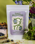 Lifestyle shot of Sweet Deliverance Blueberry & Sunflower Butter Granola (9.5 oz) bag with granola in yogurt and purple flowers and green leaves in the background