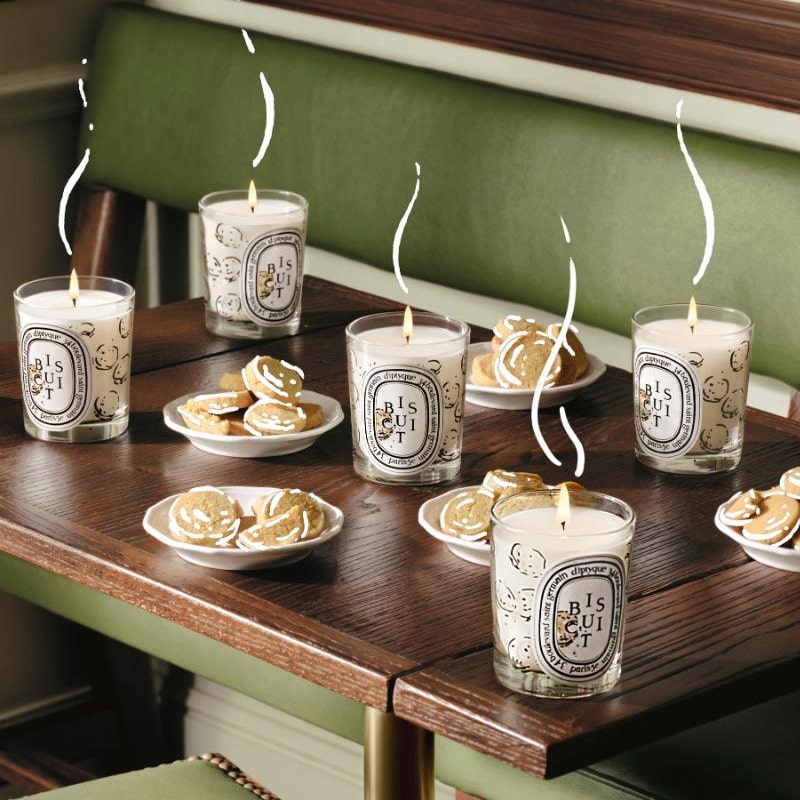 Lifestyle shot of multiple Diptyque Biscuit (Cookie) Candles (190 g) sitting on table with plates full of biscuits/cookies