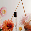 Lifestyle shot of Orchid + Ash Idyll Perfume Travel Spray - Jasmine Incense + Vanilla (10 ml) with flowers in and burning incense sticks in the background