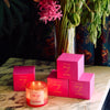 ROEN Candles De Las Flores Scented Candle - product shown lit next to boxes and flowers