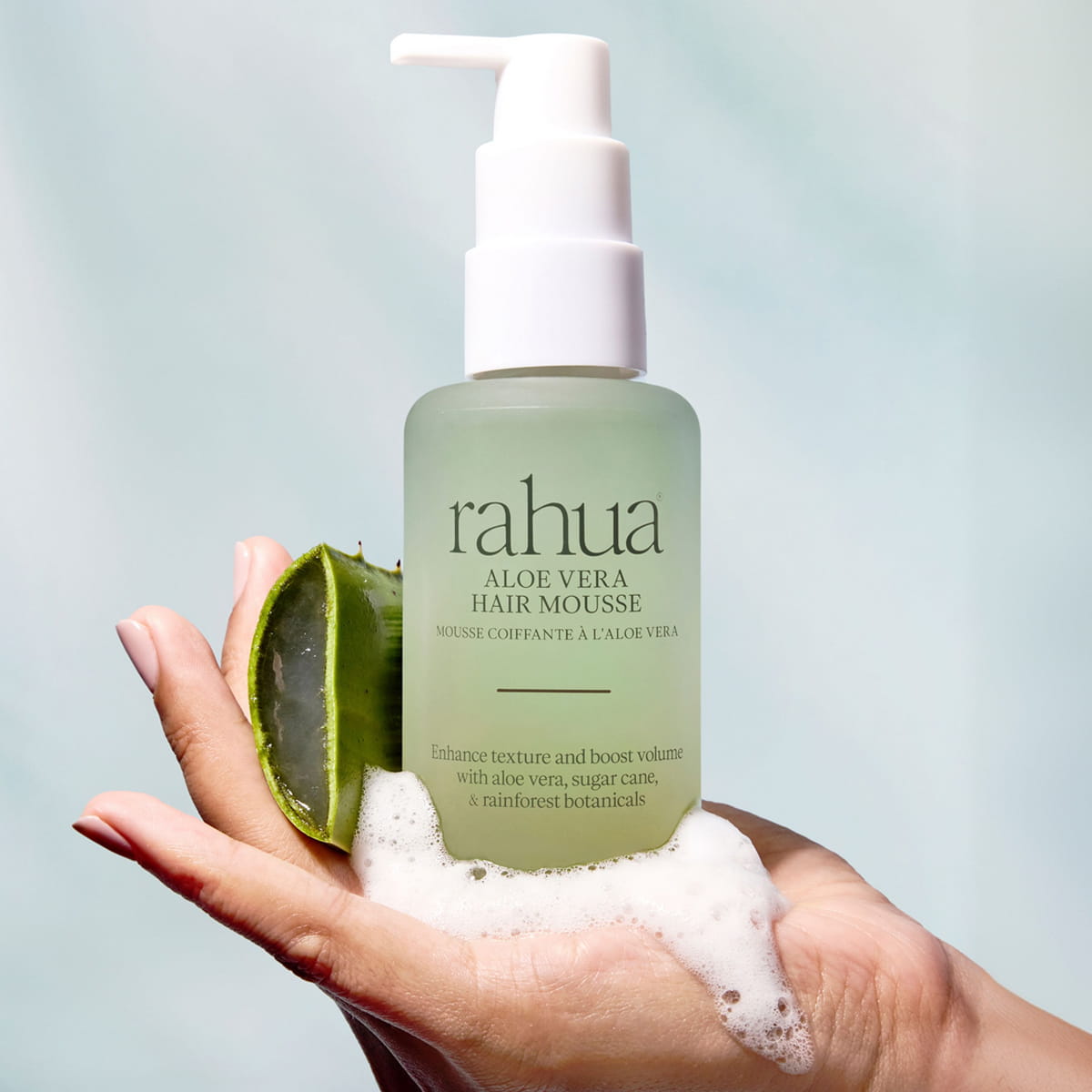 A bottle of Rahua Aloe Vera Hair Mousse (105 ml) is held in a hand with foam mousse in the palm of the hand alongside a sliced aloe vera leaf