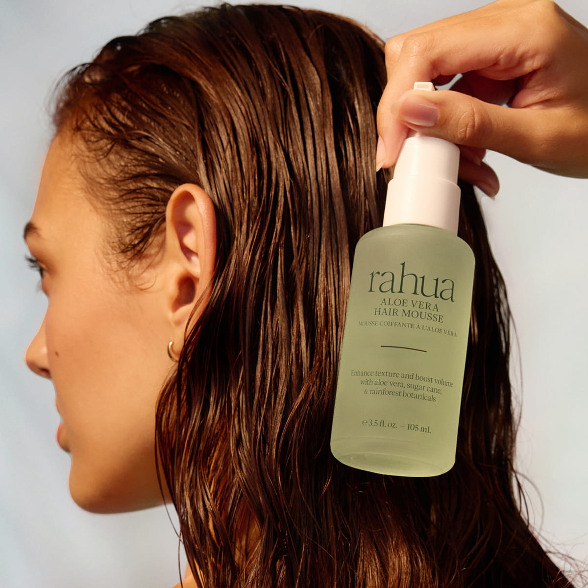 A hand holding a bottle of Rahua Aloe Vera Hair Mousse (105 ml) against a model's wet brown hair