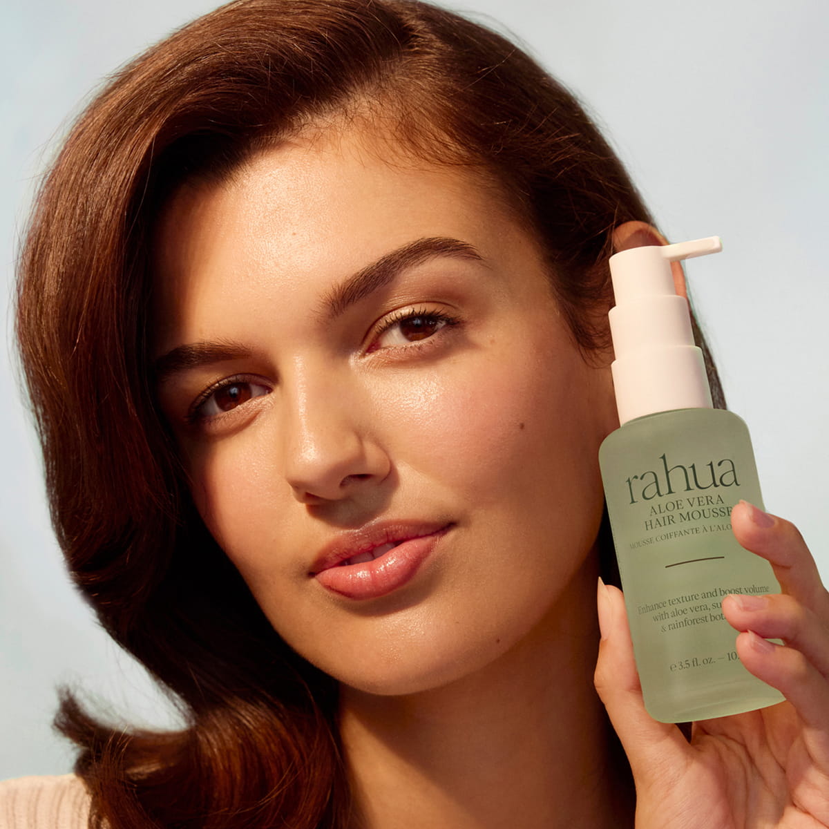 A woman with wavy brown hair holds a bottle of Rahua Aloe Vera Hair Mousse (105 ml)