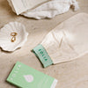 Folia Body Exfoliating Tan Removal Mitt - product shown next to towel, dish, earrings