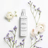 Lifestyle shot top view of Leonor Greyl Complexe Energisant - Leave-In Invigorating Scalp Treatment For Thinning Hair (150 ml) with purple flowers andn white roses placed around the bottle