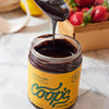 Coop’s Original Hot Fudge (10 oz) with lid off and spoon full of hot fudge dripping into jar, surrounded by strawberries and lemons.