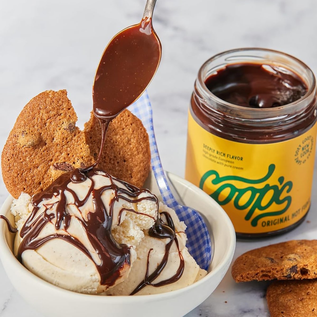 Coop’s Original Hot Fudge (10 oz) with lid off and spoon full of hot fudge drizzeld in bowl of ice cream and cookies.