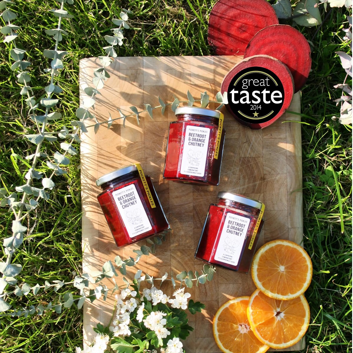 Each bottle sold individually. Puckett’s Pickles Beetroot & Orange Chutney (200 g) showing 3 bottles on wood cutting board with sliced orange and sliced beet with sprigs of eucalyptus and white flowers and grass in background. Has a seal that says great taste 2014.