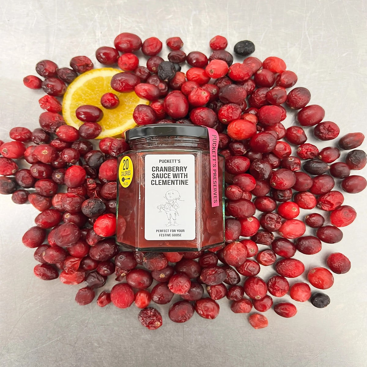 Puckett’s Pickles Cranberry, Redcurrant & Clementine (200 g) showing bottle on top of cranberries and redcurrants with a slice of lemon.