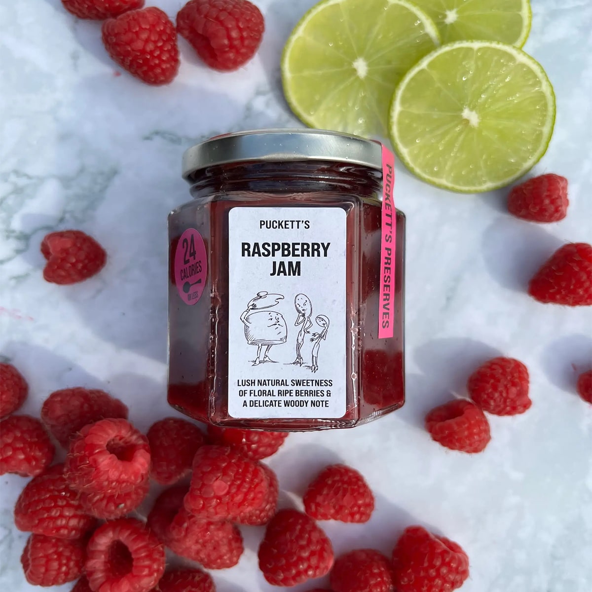 Puckett’s Pickles Raspberry Jam (226g) bottle shown with raspberry fruit and sliced lime all on marble background