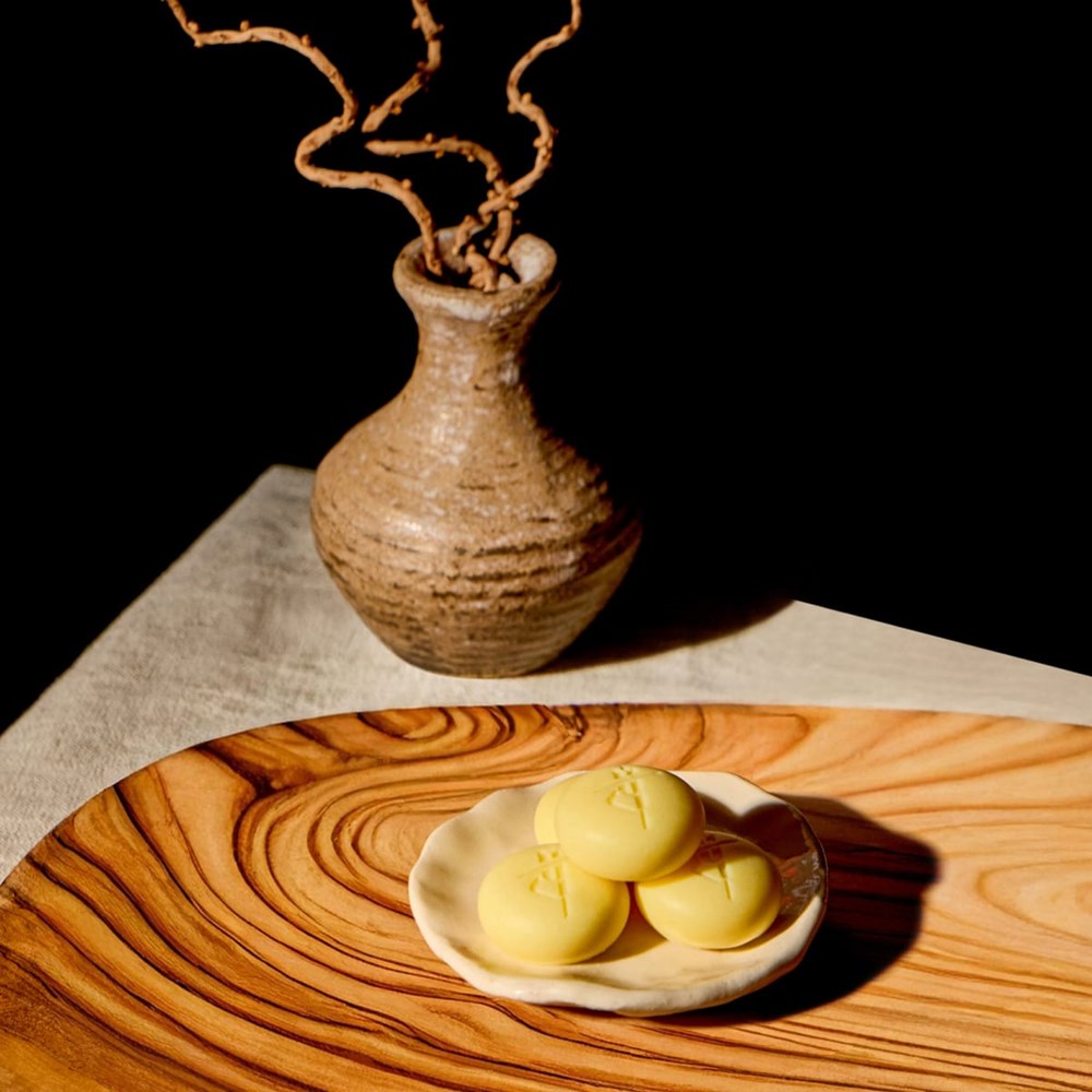 Kate McLeod Amber Vanilla Pebble Deep Moisturizing Solid Bath & Shower Oil (20 pc) - Product shown in wooden bowl with vase behind it.