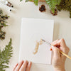 Person painting Emily Lex Studio Mushrooms Paintable Notecards (8 pcs) with greenery around.
