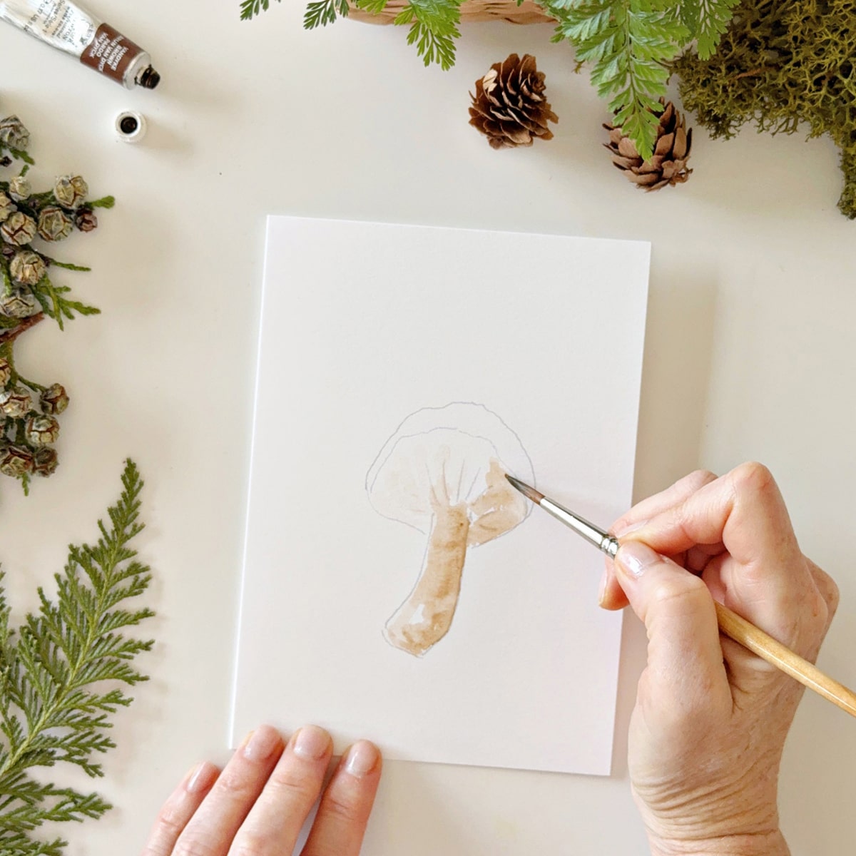 Person painting Emily Lex Studio Mushrooms Paintable Notecards (8 pcs) with greenery around.