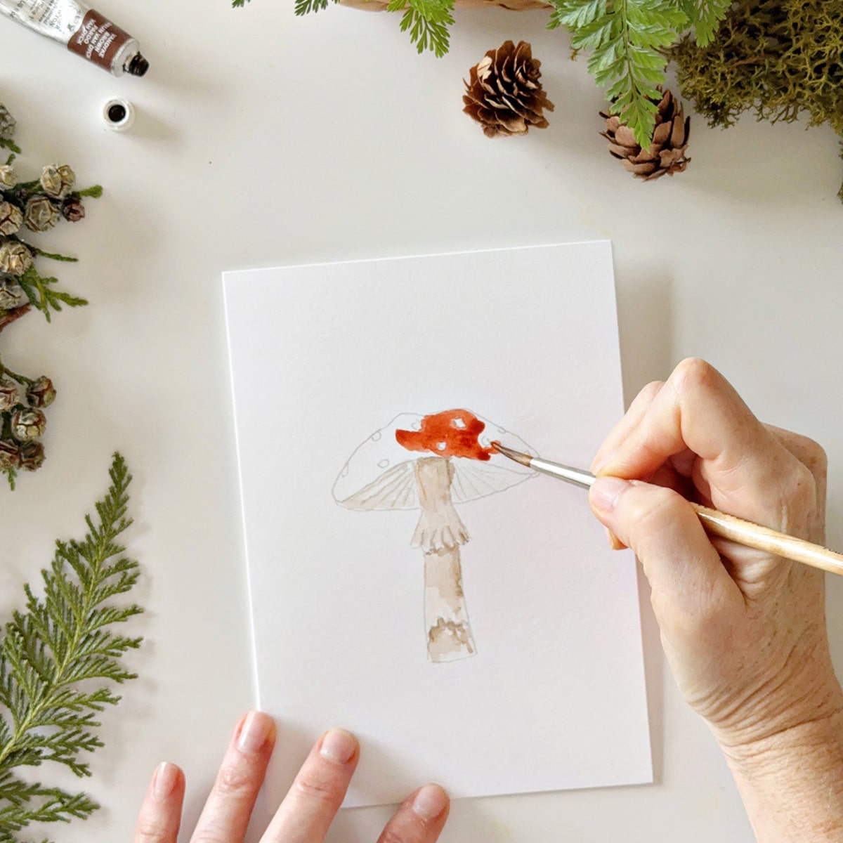 Person painting Emily Lex Studio Mushrooms Paintable Notecards (8 pcs) with greenery around.