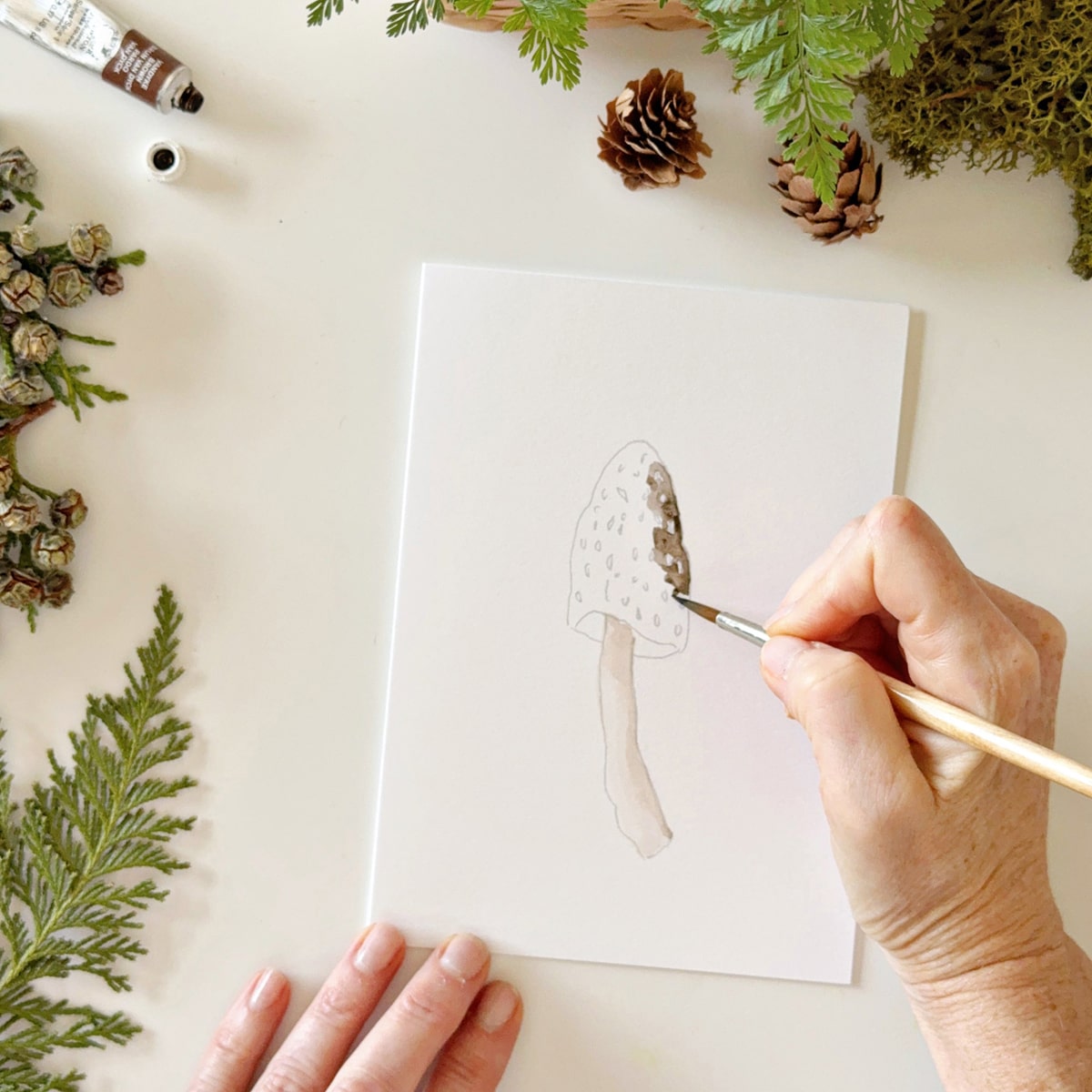 Person painting Emily Lex Studio Mushrooms Paintable Notecards (8 pcs) with greenery around.