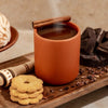 Nopalera Chocolate Ibarra Candle (8.5 oz) shown using reusable vessel as a mug with steaming hot chocolate, with cinnamon stick, cookies, and chocolate on a wooden surface.