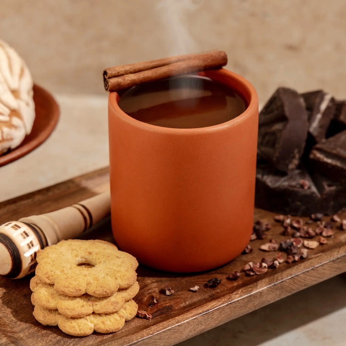 Nopalera Chocolate Ibarra Candle (8.5 oz) shown using reusable vessel as a mug with steaming hot chocolate, with cinnamon stick, cookies, and chocolate on a wooden surface.