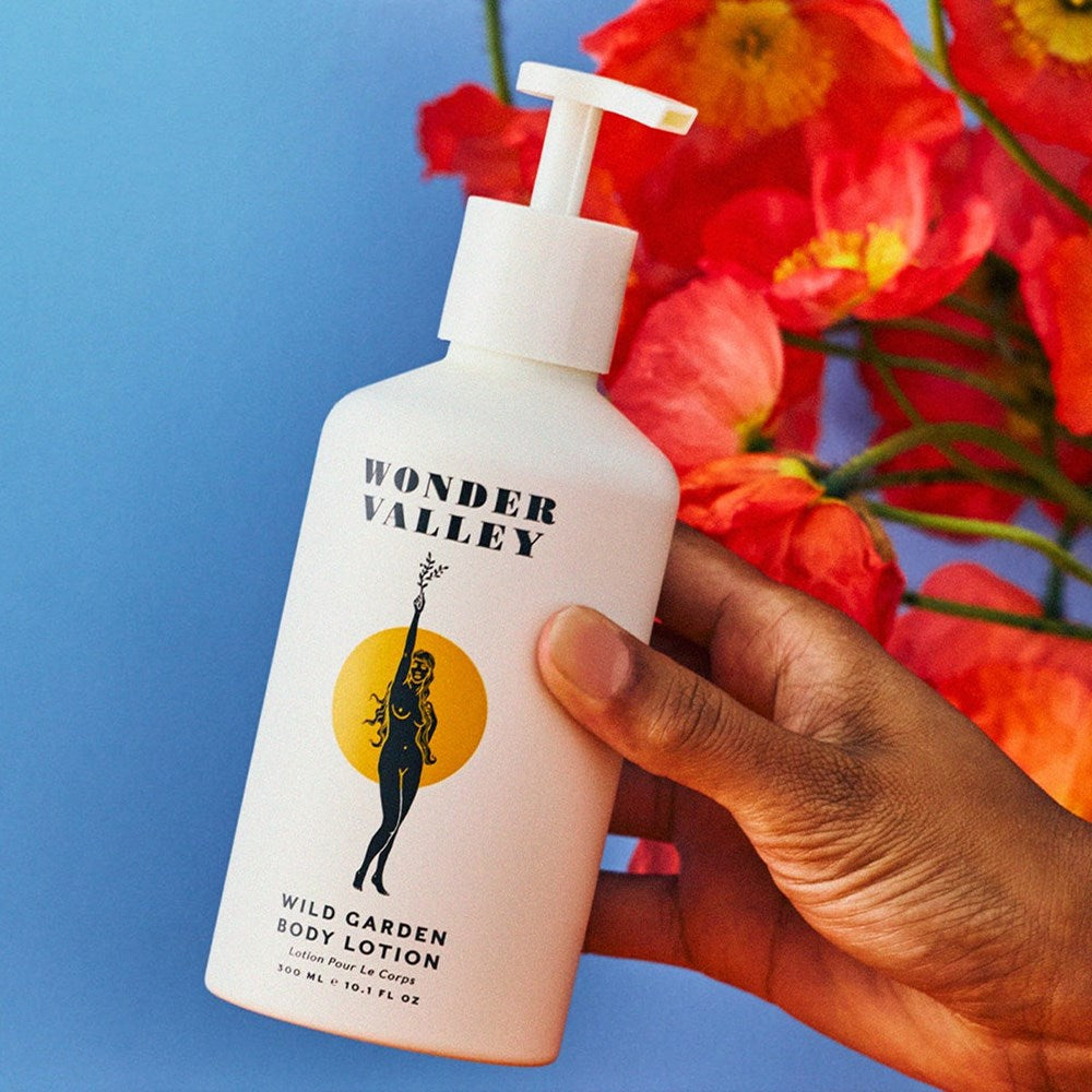 Hand holding a bottle of Wonder Valley Wild Garden Body Lotion (300 ml) against a blue background with red poppy flowers.
