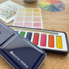 Close up of Breathe People Watercolor Paint Set on a wooden surface with color swatches.