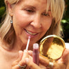 Woman applying Minori Lip Gloss - Blossom (3.5 ml) using a compact mirror outdoors