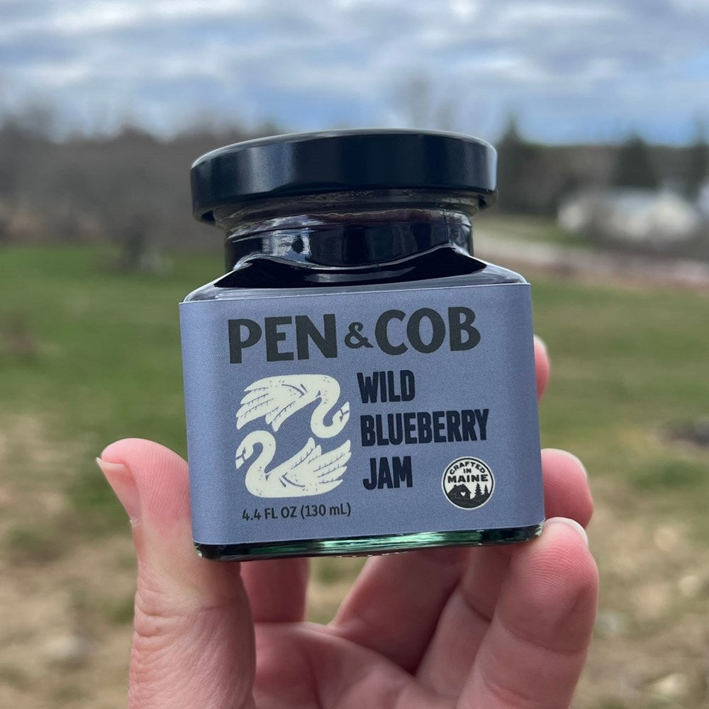 Pen & Cob Farm Wild Blueberry Jam (9 oz) - product shown being held up by hand in front of green forest area. 