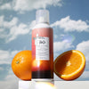 R+Co Sun Catcher Power C Styling Spray (5 oz) - product shown next to oranges and a blue sky.