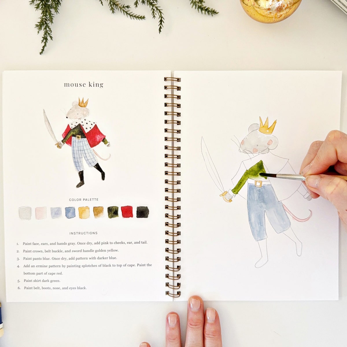 Emily Lex Studio Nutcracker Watercolor Workbook (1 pc) - page of product shown with hand and paintbrush. 