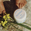 Minois Paris Magic Balm (50 ml) - product shown on ground next to feet and flowers.