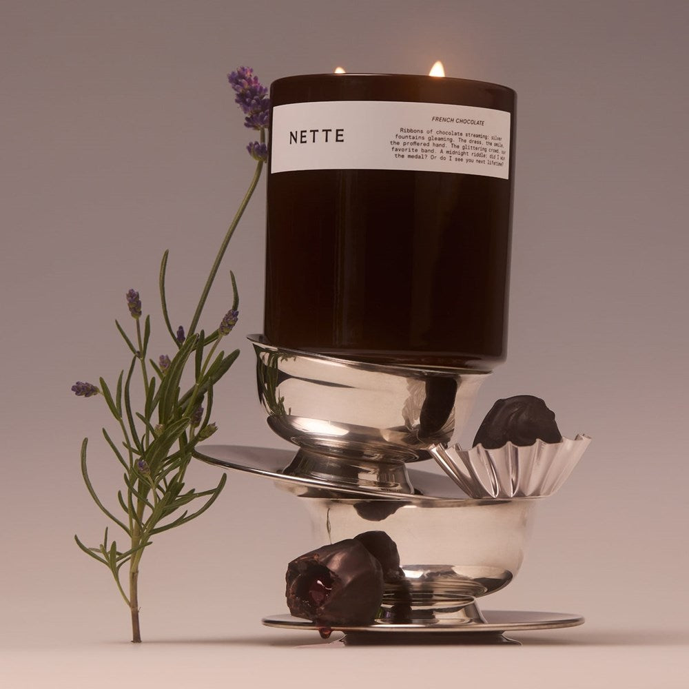 NETTE French Chocolate Scented Candle (11 oz) - product shown balancing on top of metal bowls with a flower and chocolate. 