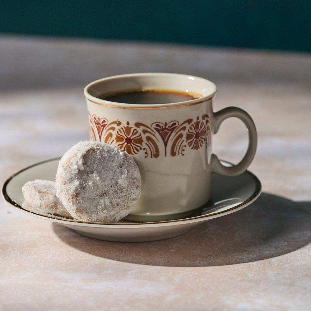 Cup of coffee with a decorative pattern on a saucer, accompanied by two Romero Luna Mexican Wedding Cookies