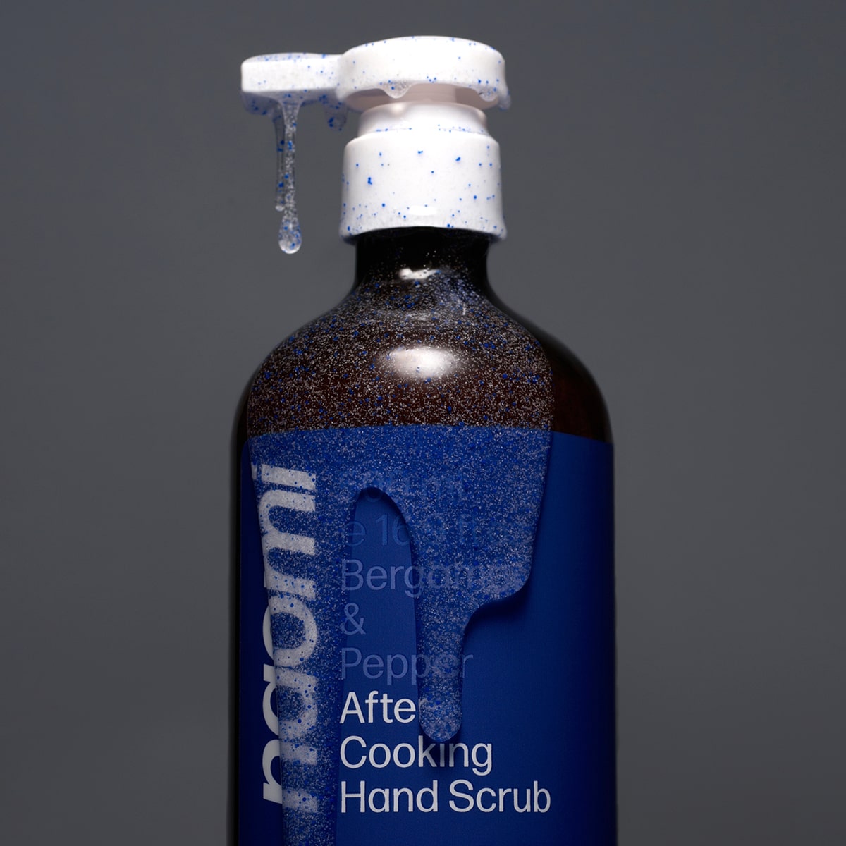 Bottle of Naomi After-Cooking Hand Scrub - Bergamot & Black Pepper Hand Wash (500 ml) with scrub coming out of pump and on bottle on a gray background
