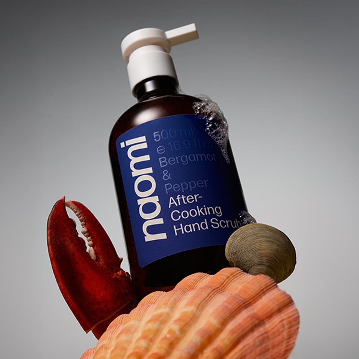 Naomi After-Cooking Hand Scrub - Bergamot & Black Pepper Hand Wash (500 ml) with a lobster claw and shellfish shells on a gray background