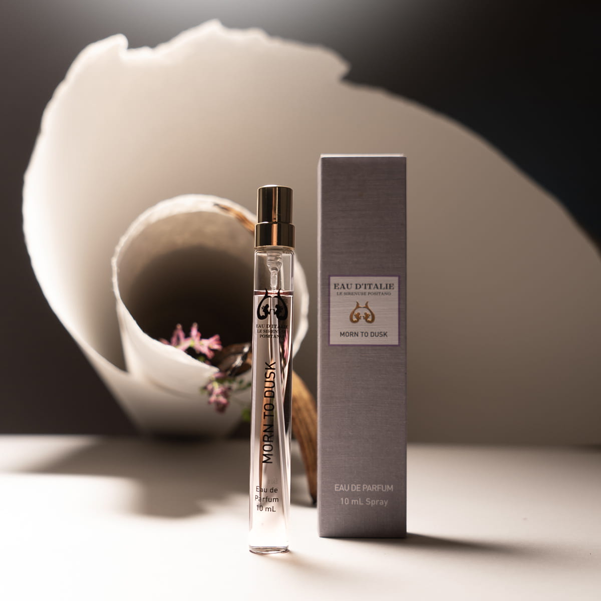 Lifestyle shot of Morn to Dusk Eau de Parfum Spray (10 ml) - product shown next to packaging with paper and flowers in the background.