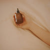 Mari Body Midsummer All-Over Oil (50 ml) - product shown being held by a hand on a sheet. 