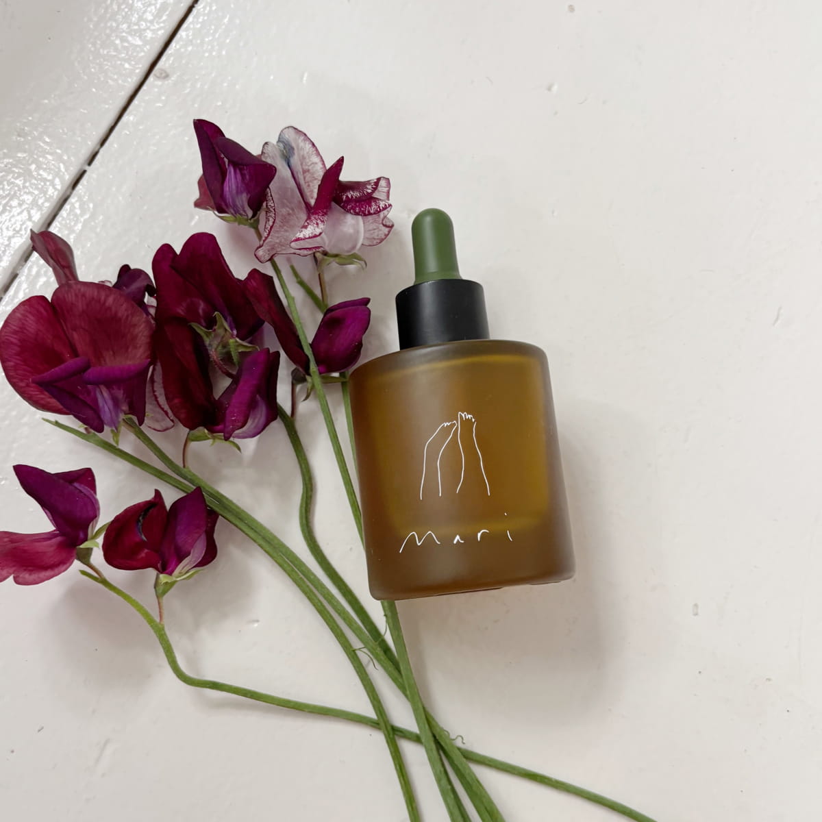 Mari Body Midsummer All-Over Oil (50 ml) - product shown on white countertop next to purple flowers. 