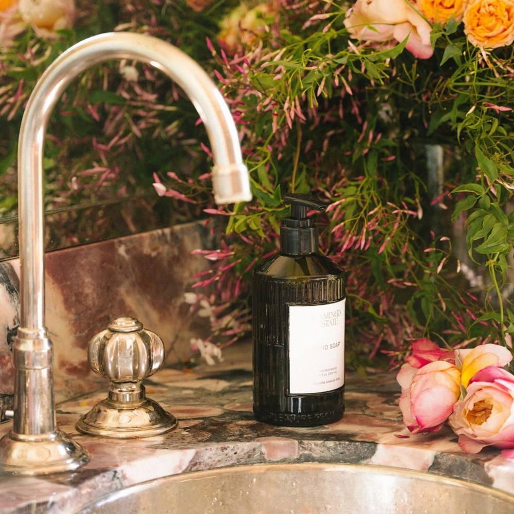 Flamingo Estate Organics Jasmine Damask Rose Glass Hand Soap - product shown on counter top next to leaves and flowers.