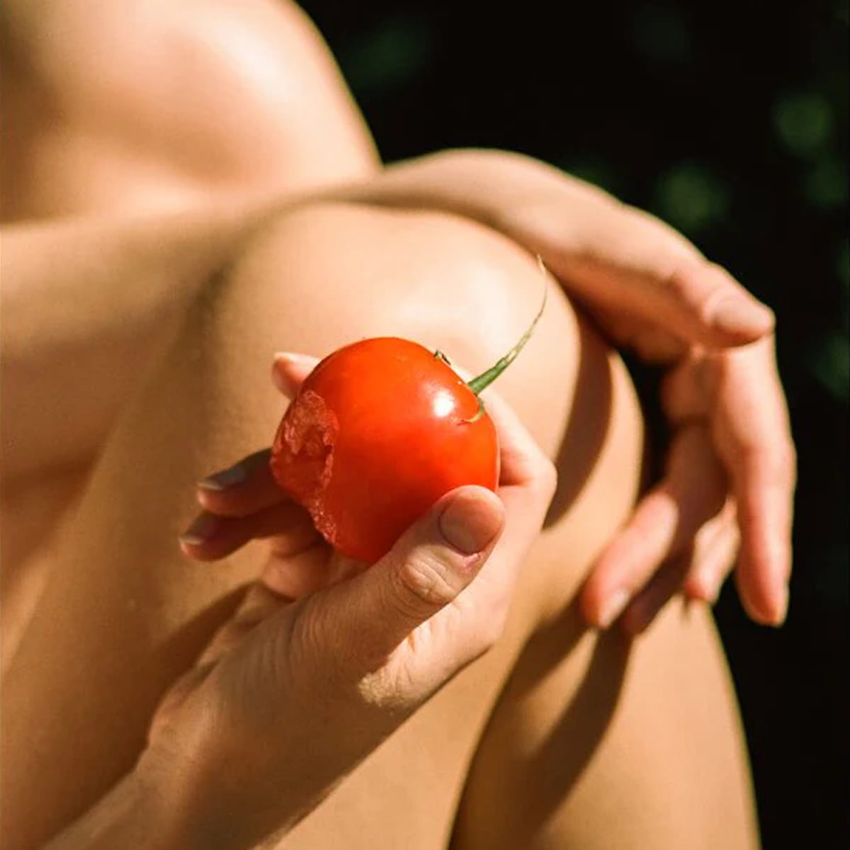 Person holding a red tomato with a bite in it against their skin outdoors
